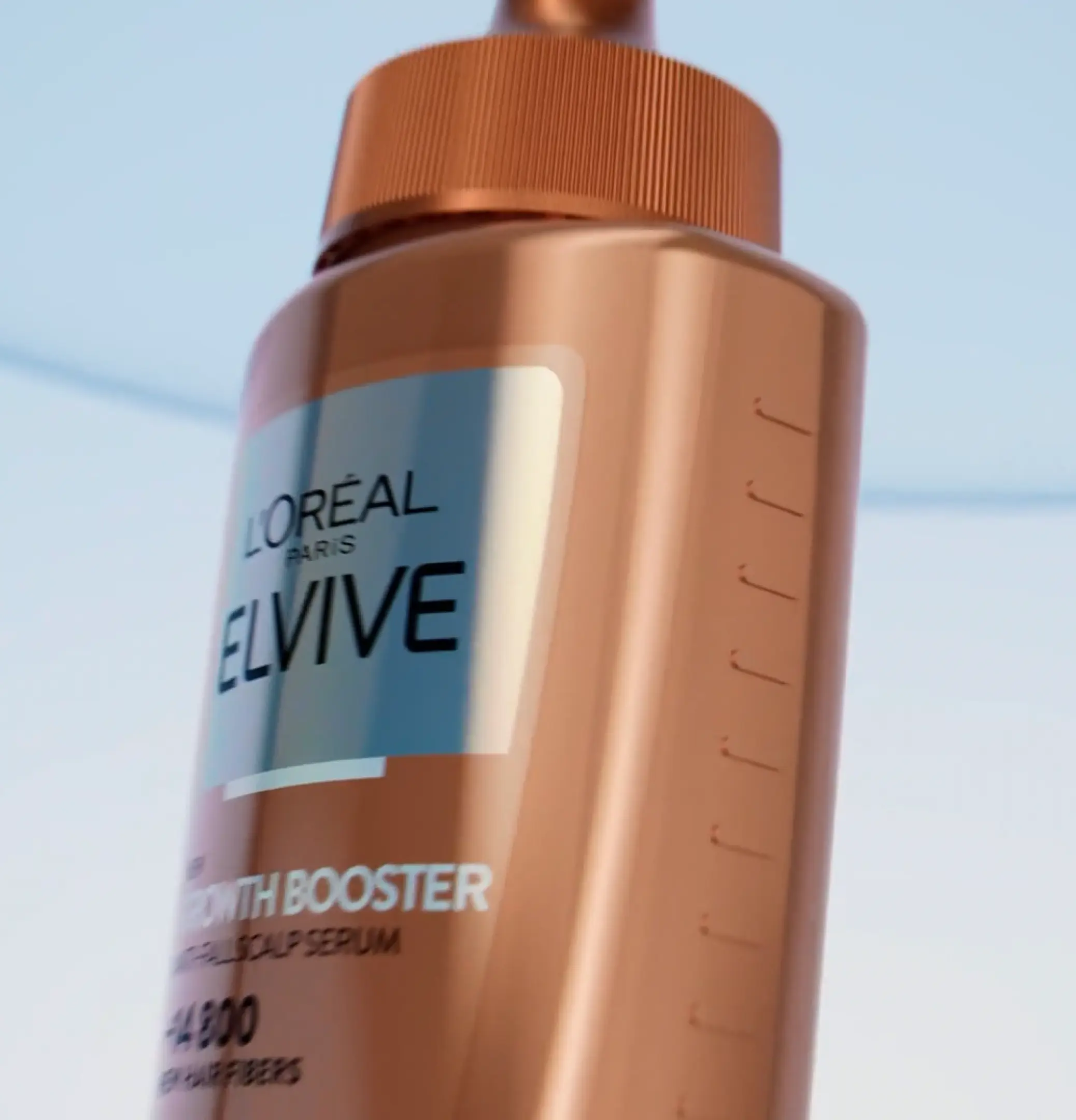 A copper L'Oreal Paris Elvive Growth Booster scalp serum bottle shot from below against a pale blue sky, its ribbed collar catching the light.